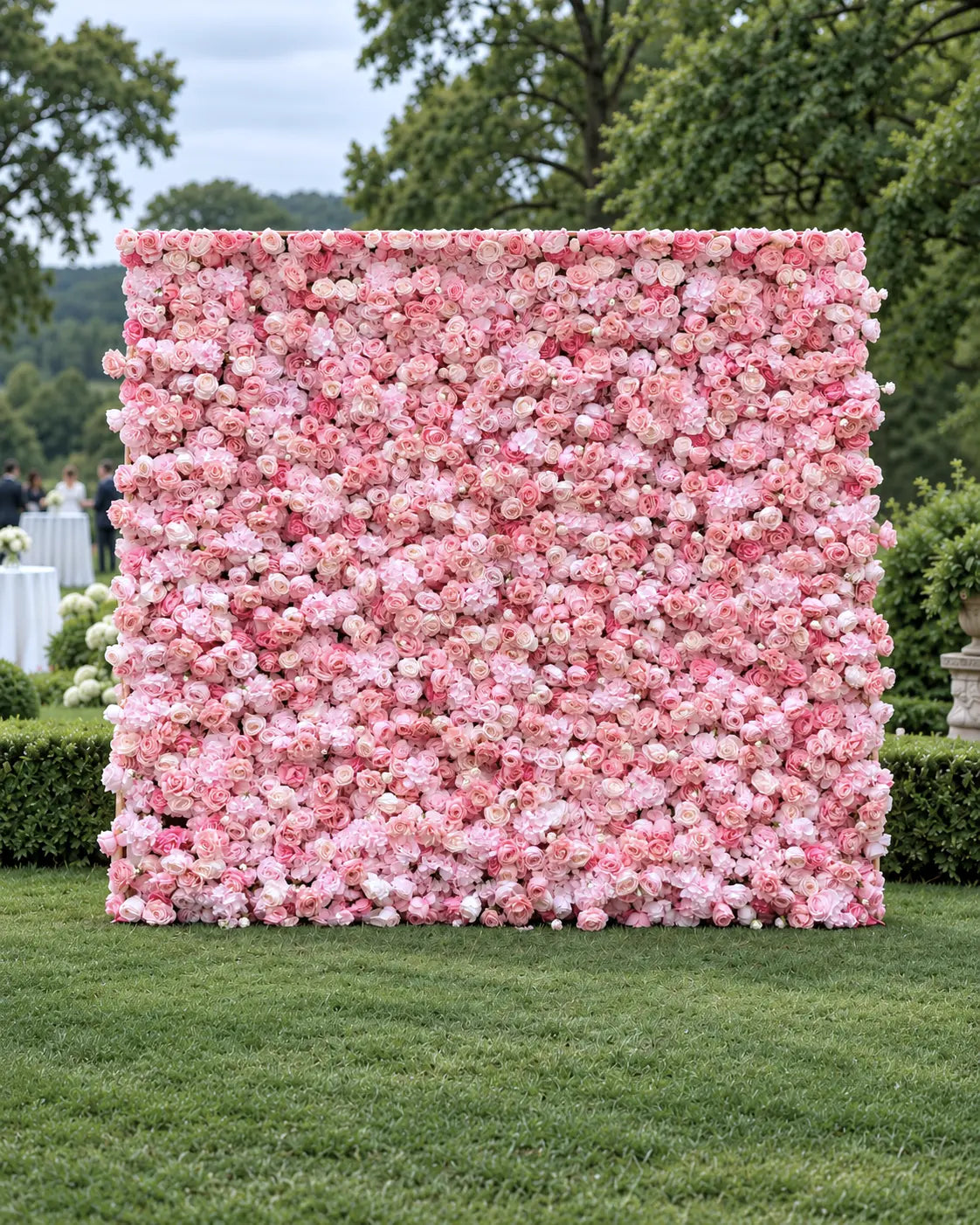 A large square floral wall backdrop covered in dense pink and white flowers, set outdoors on a green lawn with trees and hedges in the background.